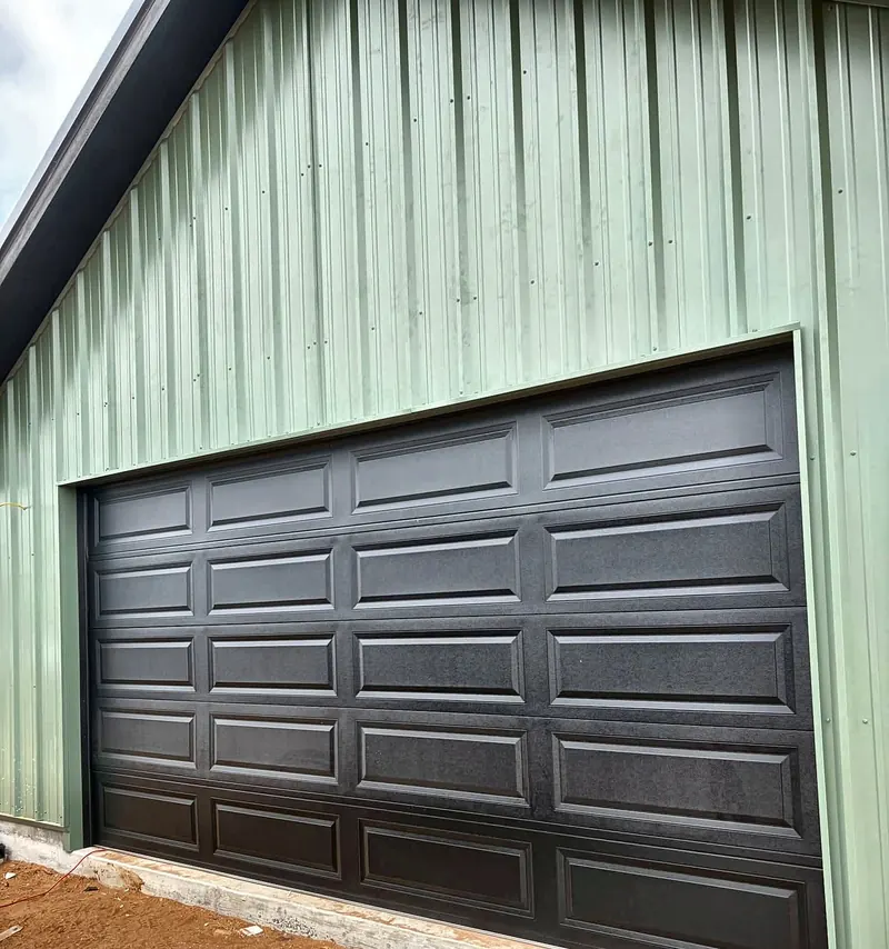 Black raised-panel garage door installed on a green metal building